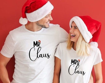 Może przedstawiać: Para w białych koszulkach z napisem "Mr. Claus" i "Mrs. Claus" czarnym pismem. Mają na sobie czapki Mikołaja i uśmiechają się do siebie.