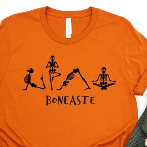 May include: Orange t-shirt with a black design of skeletons doing yoga poses. The text "BONEASTE" is below the skeletons.