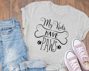 May include: A gray t-shirt with the text "My Kids Have Paws" in black lettering. The text is on a white bone shape with paw prints on either side. The shirt is laid out on a wooden surface with a pair of pink canvas shoes and a pair of blue jeans.