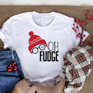 May include: White t-shirt with a red and white Santa hat, glasses, and the text "Oh Fudge". The shirt is laying on a brown burlap surface with blue jeans, brown boots, and a red and white plaid scarf.
