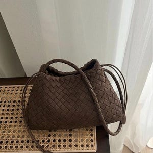 May include: A brown woven handbag with a braided handle and drawstring closure. The bag is made of a textured material and sits on a dark wooden bench with a woven top. The background includes a white curtain.
