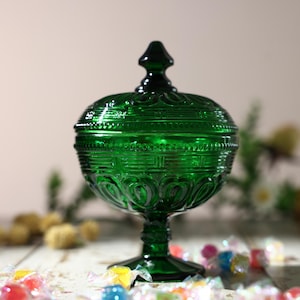 Puede incluir: Un cuenco para dulces de vidrio verde esmeralda profundo con tapa a juego. El recipiente cuenta con una base de pedestal y patrones en relieve intrincados, incluyendo bandas horizontales de puntos y líneas verticales, rematado con un remate puntiagudo.