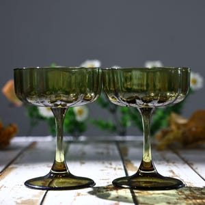Puede incluir: Un par de copas de cristal color verde oliva con un diseño de borde festoneado y un tallo delgado que termina en una base circular. La cristalería tiene una calidad translúcida con nervaduras verticales en el cuenco, creando reflejos.