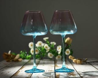 May include: Two glass wine glasses featuring a distinct ombre effect, transitioning from a soft pink hue at the top of the bowl to a vibrant turquoise blue at the base and stem. The glasses have a wide, angular bowl shape and a slender stem.