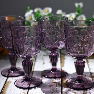 Op de afbeelding: Een set van vier paarse glazen bekers met een reliëf bloemenpatroon op de kelk. Elke beker heeft een korte, gefacetteerde steel en een ronde voet. Het glas is doorschijnend met een diepe violette tint en gedetailleerde bloemmotieven.
