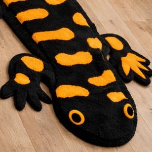 May include: A black and orange salamander-shaped rug. The rug features a black body with orange spots and a matching head and feet. The eyes are orange circles. The rug is made of a soft, plush material.