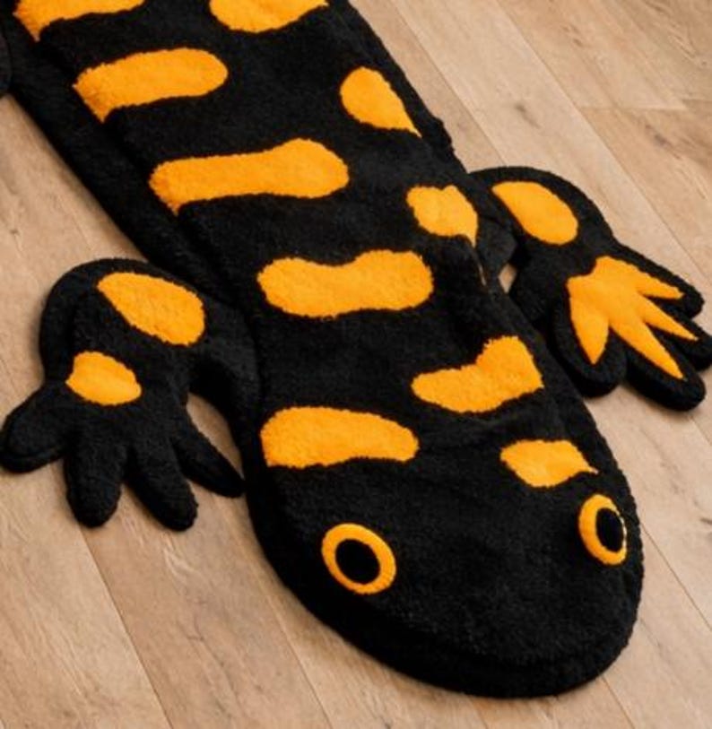 May include: A black and orange salamander-shaped rug. The rug features a black body with orange spots and a matching head and feet. The eyes are orange circles. The rug is made of a soft, plush material.