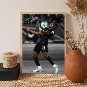 Puede incluir: Impresión en blanco y negro enmarcada de un futbolista en acción, equilibrando un balón de fútbol. El jugador lleva un uniforme negro con el número 20. La impresión incluye una firma en la esquina superior izquierda. El marco es de color madera clara.