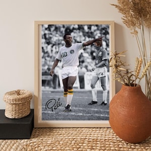 Könnte beinhalten: Gerahmtes Schwarz-Weiß-Foto von Pelé, einem Fußballspieler, der mit dem Arm zeigt. Er trägt ein weißes Trikot mit der Nummer 10 und weiße Shorts. Das Foto befindet sich in einem hellen Holzrahmen. Die Unterschrift "Pelé" ist sichtbar.