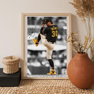 May include: A framed black and white photograph of a baseball pitcher in a black jersey with yellow lettering reading SKENES and the number 30. The player wears white trousers, yellow cleats, and a black cap, captured in a pitching motion.