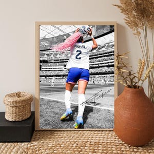 May include: Framed black and white print of a soccer player in a stadium. The player, wearing a white jersey with the number 2 and blue shorts, is throwing a soccer ball. The player has pink hair. The print is in a light brown frame.