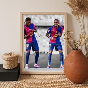 May include: Framed print featuring two soccer players in blue and pink jerseys, numbered 19 and 11. The players are in a stadium setting, with a black and white background. The print is in a light wood frame.