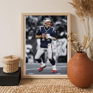 Op de afbeelding: Ingelijste print met een American footballspeler in een marineblauw en wit uniform, die een bal vasthoudt. De speler draagt een helm en rode schoenen. De achtergrond is in zwart-wit. De print zit in een licht houten frame.