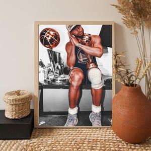 Pode incluir: Fotografia emoldurada de um jogador de basquete com uma camisa azul e dourada, segurando um troféu e uma bola de basquete. A imagem está em uma moldura de madeira, com um vaso decorativo e uma cesta em primeiro plano.
