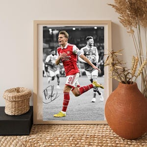 May include: A framed black and white photograph of a soccer player in a red and white jersey with the number 8, running on a field. The player wears bright yellow cleats and has a black ink signature on the lower left side of the print.