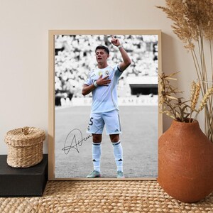 May include: A framed black and white photograph of a soccer player in a white uniform, number 5, with a signature at the bottom. The player is standing on a field, raising one arm and touching his chest with the other.
