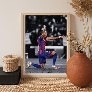 May include: Framed print featuring a soccer player in a blue and red uniform, kneeling on one knee with arms raised in celebration. The background is in black and white, with the player's signature at the bottom. The frame is light brown.