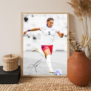May include: Framed print of a soccer player in a white jersey with the number 5 and "USA" on the chest, kicking a soccer ball. The player wears red shorts and white socks. The print includes a signature and is set against a black and white background.