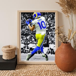 Puede incluir: Impresión enmarcada de un jugador de fútbol americano con uniforme blanco, azul y amarillo atrapando un balón. La camiseta del jugador tiene el número 10. El fondo es una imagen en blanco y negro de la multitud. El marco es de color marrón claro.