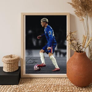 Könnte beinhalten: Gerahmter Druck eines Fußballspielers in blauem Trikot, der einen Fußball tritt. Der Spieler trägt weiße Socken und rote Schuhe. Der Druck hat einen hellen Holzrahmen und eine Signatur auf dem Ball.