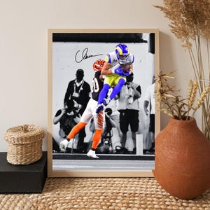 以下が含まれることがあります： 青と黄色のユニフォームを着たフットボール選手が空中キャッチをしているフレーム入りのプリント。画像は白黒で、選手のユニフォームはカラーです。サインが見えます。フレームは明るい木の色です。