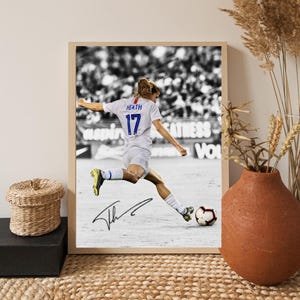 May include: Framed black and white photograph of a soccer player kicking a soccer ball. The player is wearing a white jersey with the name "HEATH" and the number "17". The photograph is signed. The frame is a light wood color.