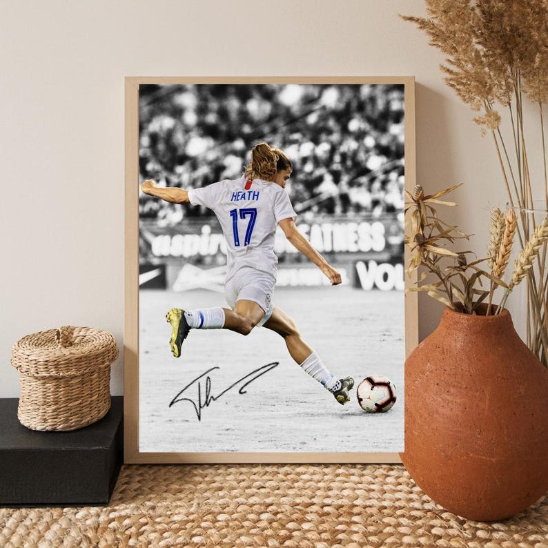 May include: Framed black and white photograph of a soccer player kicking a soccer ball. The player is wearing a white jersey with the name "HEATH" and the number "17". The photograph is signed. The frame is a light wood color.