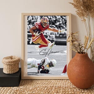 May include: Framed print featuring a football player in a red and gold uniform leaping with the ball. The background is black and white, with a signature at the bottom. The frame is light brown.