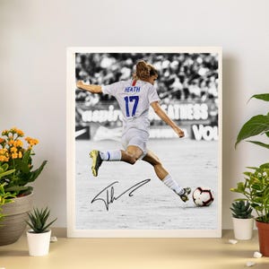May include: Framed black and white photograph of a soccer player in a white uniform with the number 17 and the name "Heath" on the back, kicking a soccer ball. The photograph is signed and displayed in a white frame.