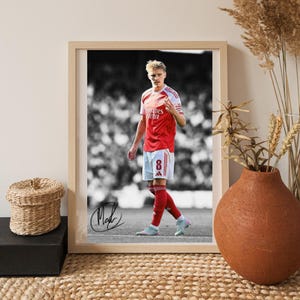May include: Framed photograph of a soccer player in a red and white jersey with the number 8. The player is standing on a green field, with a black and white background. The photograph is signed in black ink.