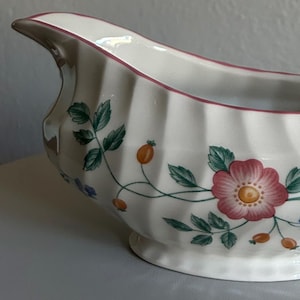 May include: White ceramic gravy boat with a floral design featuring pink flowers, green leaves, and orange berries. The rim is trimmed with a pink border. The boat has a curved handle and a spout.