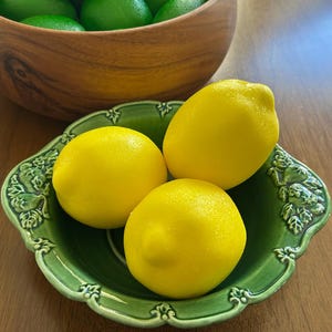 May include: Three bright yellow lemons rest in a decorative green ceramic dish with ornate edges. A wooden bowl filled with green limes is in the background. The lemons are the focal point, showcasing their vibrant color and texture.