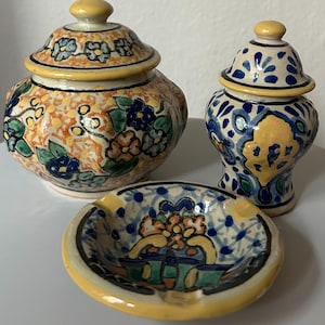 May include: Three pieces of hand-painted ceramic pottery. The set includes a small round ashtray, a small jar with a lid, and a taller jar with a lid. The pieces are decorated with floral patterns in orange, blue, green, and yellow.