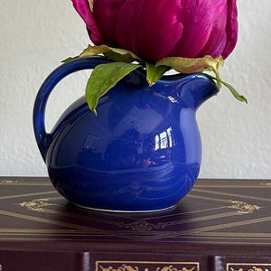May include: A vibrant magenta peony flower in a small, cobalt blue pitcher. The pitcher has a curved handle and a spout. The flower is the focal point, with green leaves peeking out. The pitcher sits on a dark, decorative book.