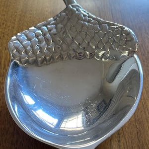 May include: A silver-colored, acorn-shaped dish. The dish has a textured top, resembling the cap of an acorn, and a smooth, concave bowl. The stem of the acorn extends upwards. The dish is likely made of metal and is designed for decorative or functional use.