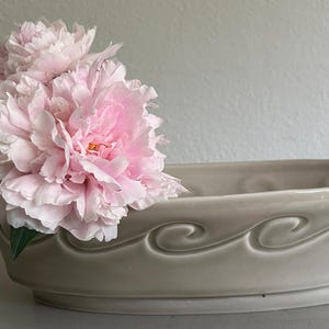 May include: An oval-shaped ceramic bowl in a light beige color featuring a raised, embossed wave pattern along the exterior side. A cluster of soft pink peony flowers with layered petals is tucked into one end of the bowl, partially obscuring the rim.