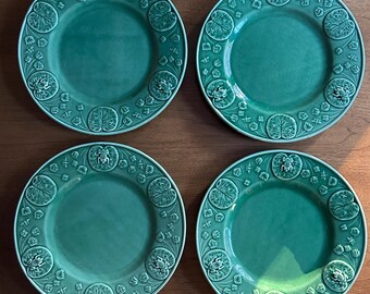 May include: Four green ceramic plates with an embossed design. Each plate has a smooth central area and a border with a repeating pattern of lily pads and frogs. The plates are displayed on a wooden surface.