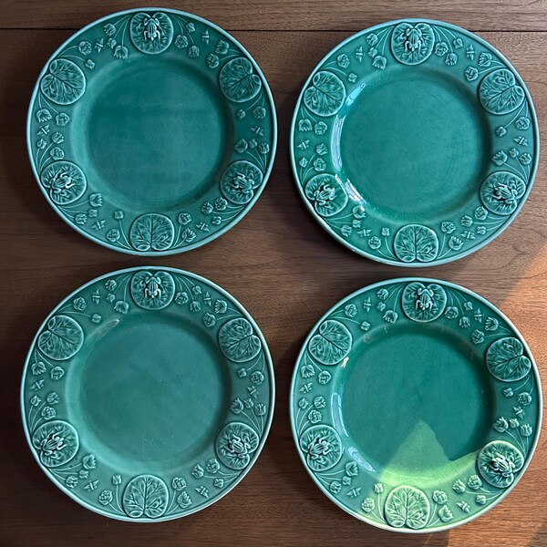 May include: Four green ceramic plates with an embossed design. Each plate has a smooth central area and a border with a repeating pattern of lily pads and frogs. The plates are displayed on a wooden surface.