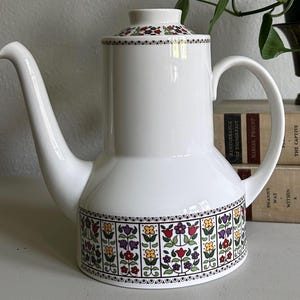 May include: White ceramic coffee pot with a floral design around the base and on the lid. The pot has a long spout and a curved handle. The floral pattern includes red, yellow, purple, and green flowers. The pot is sitting on a white surface.