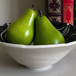 May include: A white ceramic bowl with a black and white patterned rim holds two green pears. The pears have a textured surface and brown stems. Decorative books are in the background.