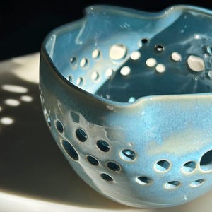 May include: A light blue ceramic bowl with a textured surface and numerous circular openings. The bowl has an irregular, wavy rim and sits on a white surface. The bowl's color is a mix of light blue and white.