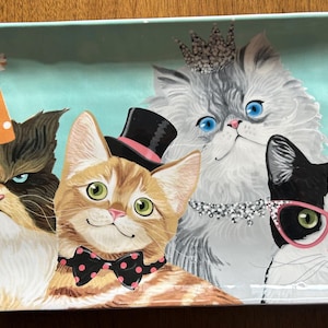 May include: Rectangular ceramic tray with a light blue background featuring four illustrated cats. One cat wears a party hat, another a top hat and bow tie, a third a crown, and the last wears pink glasses. The cats have various fur colors.