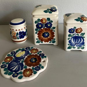 May include: A set of four ceramic kitchen items featuring hand-painted floral patterns in blue, brown, and green on an off-white background. The collection includes two square canisters with lids, one cylindrical jar with a lid, and a flat, scalloped trivet.