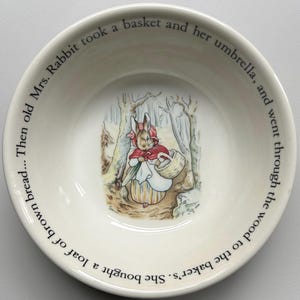 May include: A cream-colored ceramic bowl featuring an illustration of Mrs. Rabbit in a red cloak and holding a basket. The bowl has text around the rim that reads, "Then old Mrs. Rabbit took a basket and her umbrella, and went through the wood to the baker's. She bought a loaf of brown bread..."