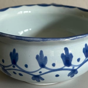 May include: A white ceramic bowl with a scalloped rim and a blue floral design. The bowl has a rounded shape and a smooth, glossy finish. The blue design features stylized leaves and vines.