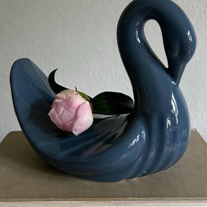 May include: A glossy, dark blue ceramic swan figurine with a curved neck and a smooth, rounded body. A single, light pink peony flower with dark green leaves is nestled within the hollow center of the swan's back, resting against the curved surface.