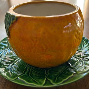 May include: A ceramic orange-shaped planter with a textured surface, resting on a green leaf-patterned saucer. The planter is a vibrant orange color with green accents, creating a naturalistic design. This decorative piece is suitable for small plants or as a unique home accent.