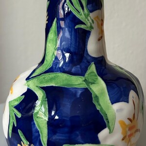 Puede incluir: Un jarr&oacute;n de cer&aacute;mica con cuerpo bulboso y cuello estrecho. El jarr&oacute;n est&aacute; pintado con un fondo azul oscuro y presenta flores blancas, hojas verdes y detalles amarillos. El jarr&oacute;n mide aproximadamente 20 cm de altura.