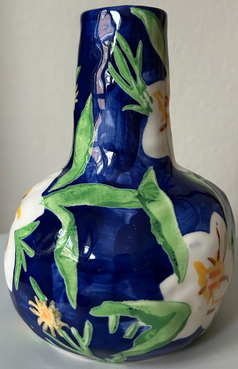 Puede incluir: Un jarr&oacute;n de cer&aacute;mica con cuerpo bulboso y cuello estrecho. El jarr&oacute;n est&aacute; pintado con un fondo azul oscuro y presenta flores blancas, hojas verdes y detalles amarillos. El jarr&oacute;n mide aproximadamente 20 cm de altura.