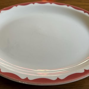 May include: White oval serving platter with a scalloped coral-colored border. The plate has a simple, elegant design, suitable for serving appetizers or main courses. The plate is made of ceramic.